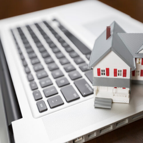 The photo features a miniature model of a two-story house with white siding and red shutters, placed on the keyboard of a laptop computer. The house has a gray roof, a small front porch, and two chimneys, resembling a typical suburban home. The laptop is silver and has a black keyboard with visible ports on the side, indicating that it may be a model used for business or professional purposes. This setup may symbolize the intersection of real estate and technology, likely used by the Randall School of Real Estate to illustrate the modern aspects of real estate education or the use of technology in the real estate industry. The focus is on the house model, suggesting a primary emphasis on residential real estate.