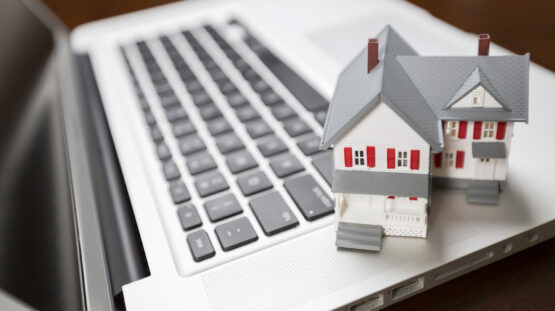 The photo features a miniature model of a two-story house with white siding and red shutters, placed on the keyboard of a laptop computer. The house has a gray roof, a small front porch, and two chimneys, resembling a typical suburban home. The laptop is silver and has a black keyboard with visible ports on the side, indicating that it may be a model used for business or professional purposes. This setup may symbolize the intersection of real estate and technology, likely used by the Randall School of Real Estate to illustrate the modern aspects of real estate education or the use of technology in the real estate industry. The focus is on the house model, suggesting a primary emphasis on residential real estate.